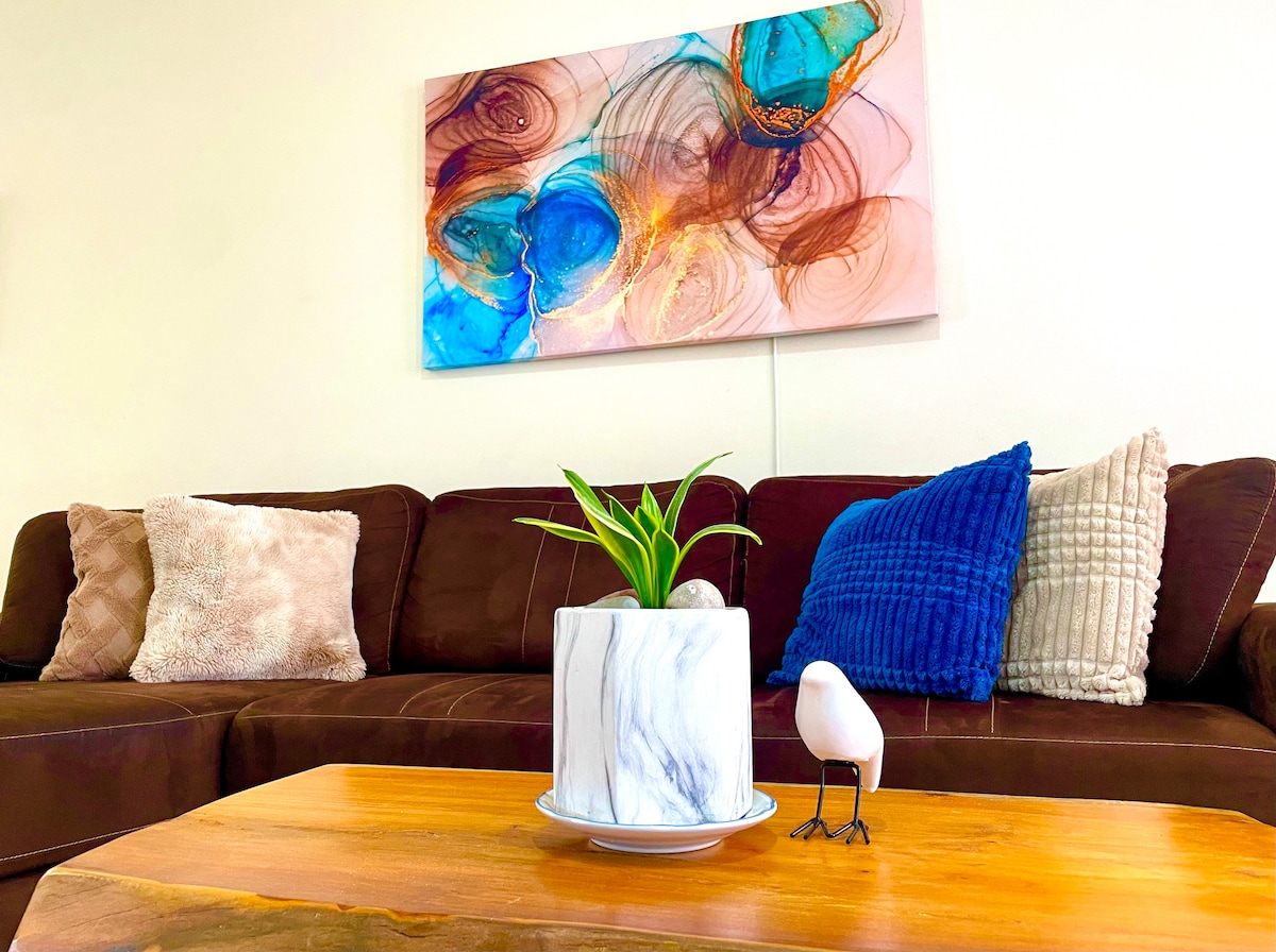 A living room scene showcases a dark brown sofa adorned with soft cushions in neutral tones and a blue textured throw. A small potted plant is placed on a round marble table, complemented by an artistic wall hanging featuring flowing colors.