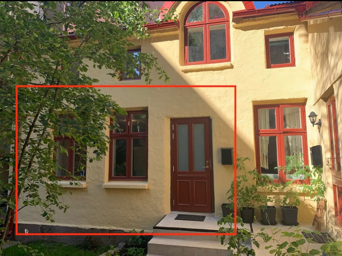 The exterior of a traditional house features soft yellow walls with red and white window frames. Large windows allow natural light, and foliage provides a touch of nature. A small entrance area leads into the home, and elements of the garden are visible, adding to the inviting atmosphere.