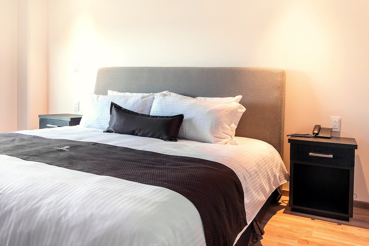 A spacious bed is positioned against a light-colored wall, with a gray upholstered headboard and crisp white linens. Dark bedside tables flank the bed, each featuring a small lamp. Natural light illuminates the room, enhancing the clean and modern ambiance.