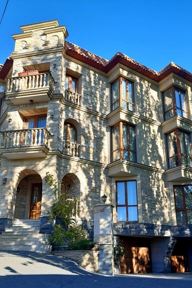 A vintage-style building features a stone façade, with multiple balconies and large windows allowing ample natural light. The entrance is marked by a wooden door, surrounded by neatly arranged greenery. The structure is set against a clear blue sky.