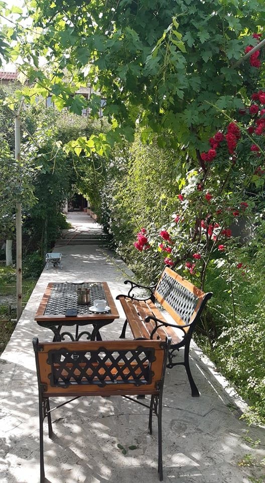 A serene garden pathway is bordered by lush greenery and vibrant rose bushes. A wooden bench with a wrought iron frame is positioned beside a small table, both set on a stone surface. Natural light filters through the surrounding foliage, creating a peaceful outdoor retreat.