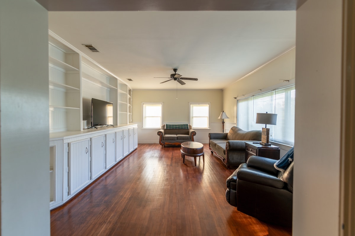 The living area is characterized by hardwood flooring and a ceiling fan. A sectional sofa and armchair are positioned near large windows, allowing for natural light. Built-in shelving units and a television are visible along one wall, enhancing the open and inviting feel.
