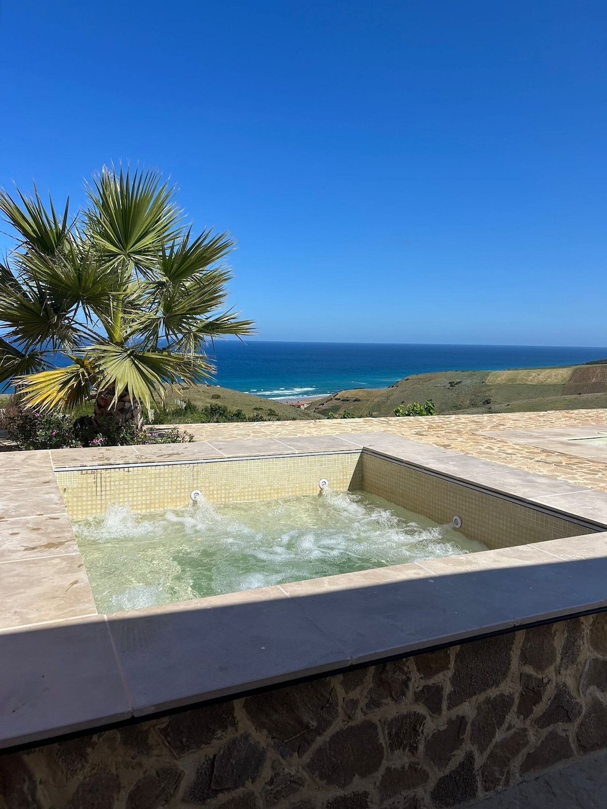 A jacuzzi is set against a backdrop of the ocean and rolling hills. The bubbling water reflects the clear blue sky. Green palm leaves add a touch of nature to the scene, enhancing the serene environment designed for relaxation.