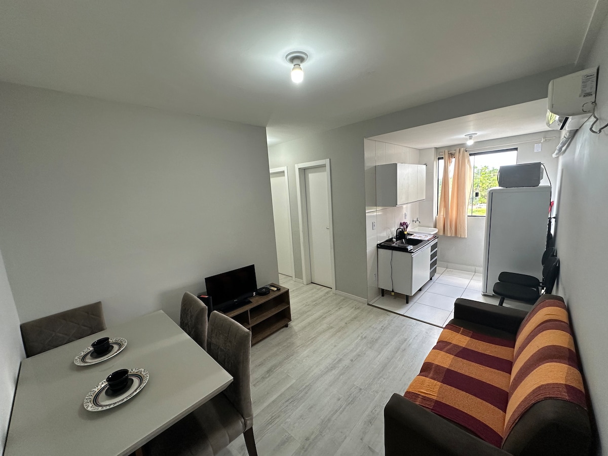 An airy living space features a dining table with four chairs and a comfortable seating area. A kitchenette is visible, equipped with essential appliances. Natural light enters through the window, enhancing the bright atmosphere of the neutral-toned decor.