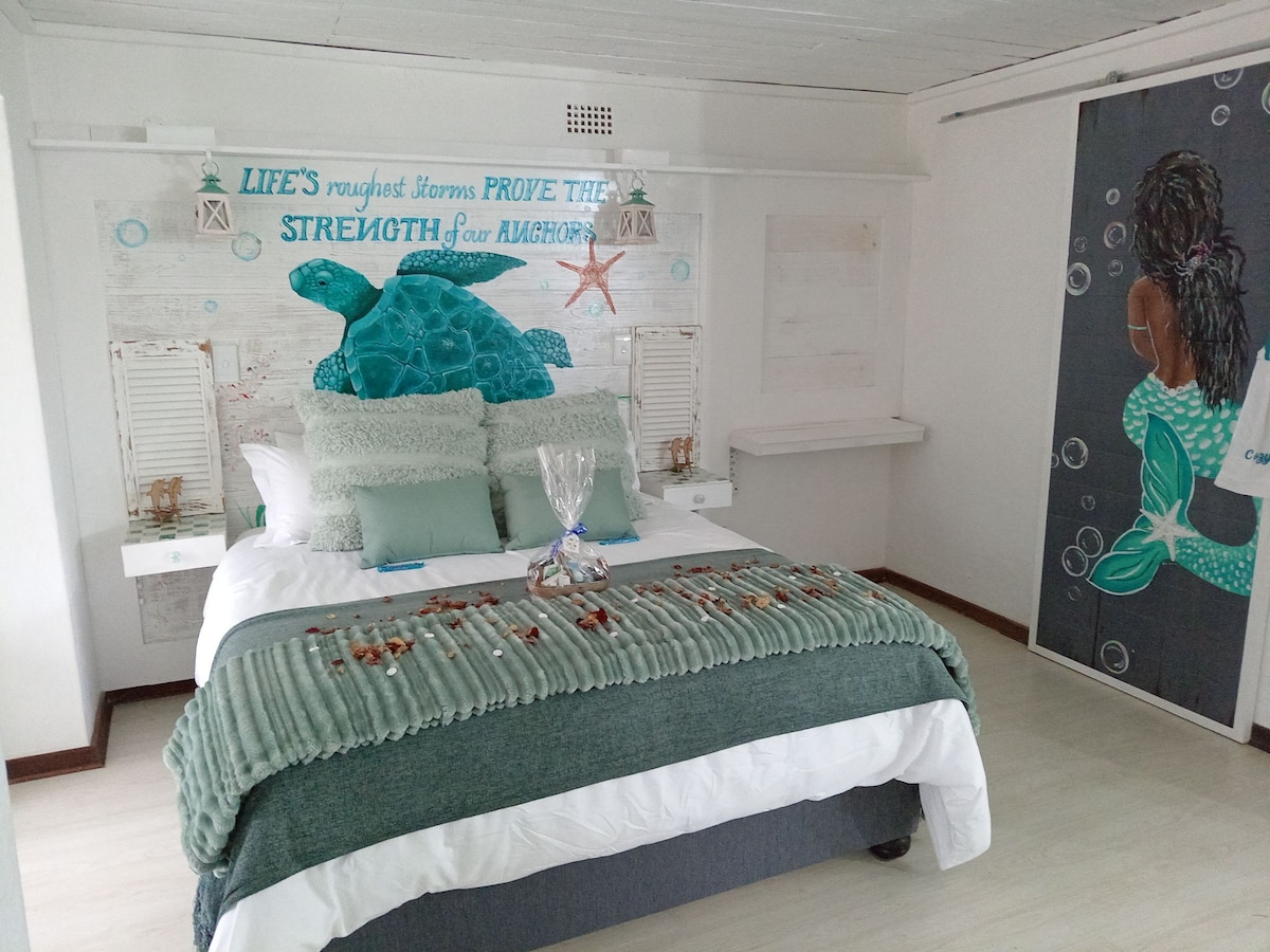 A serene bedroom features a spacious bed adorned with a soft, textured blanket and decorative pillows. A nautical-themed wall art with a turtle is displayed above the bed. Light-colored walls and floor enhance the calming ambiance, while a mural depicts a figure by the ocean.