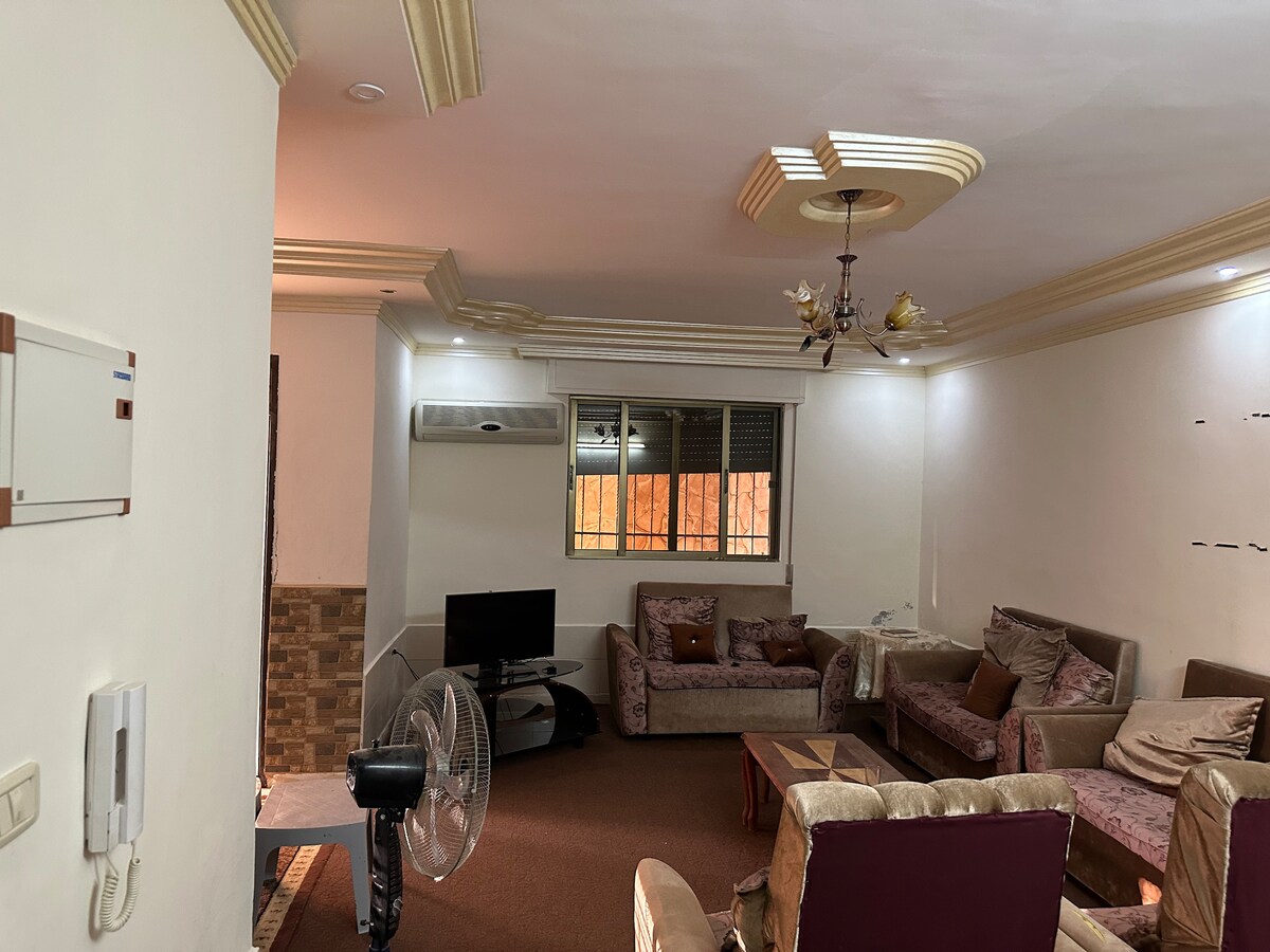 A spacious living area is furnished with a combination of sofas and armchairs. Natural light filters through a large window. A television is positioned on a stand, and a ceiling fan is visible on one side, providing comfort in the room.