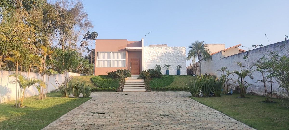 A modern house is presented with a textured façade and a large front entrance. A pathway of stone leads to the door, bordered by well-maintained grass and tropical plants. The clear sky above creates a serene atmosphere, enhancing the inviting exterior.