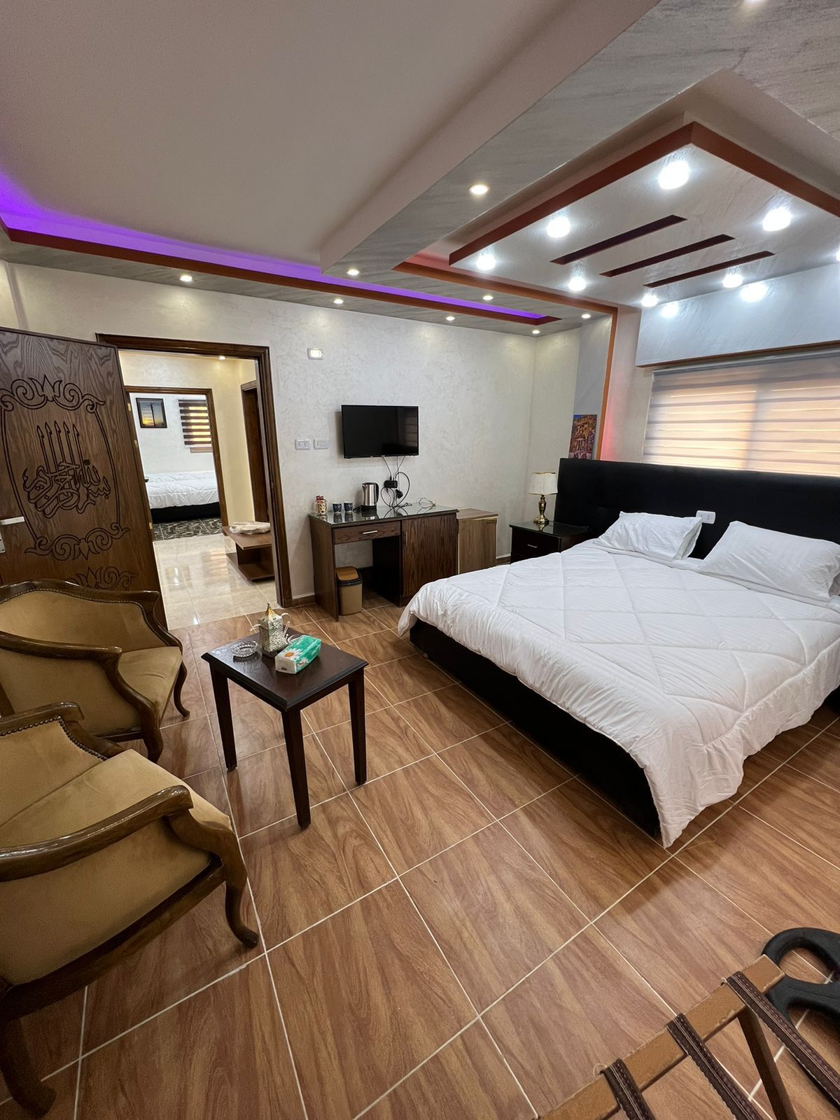 The room features a spacious bed adorned with a crisp white comforter. Two elegant chairs are positioned beside a small table. A flat-screen TV is mounted on the wall, and soft lighting is provided by recessed ceiling fixtures. A warm atmosphere is created by the wooden door and furnishings.