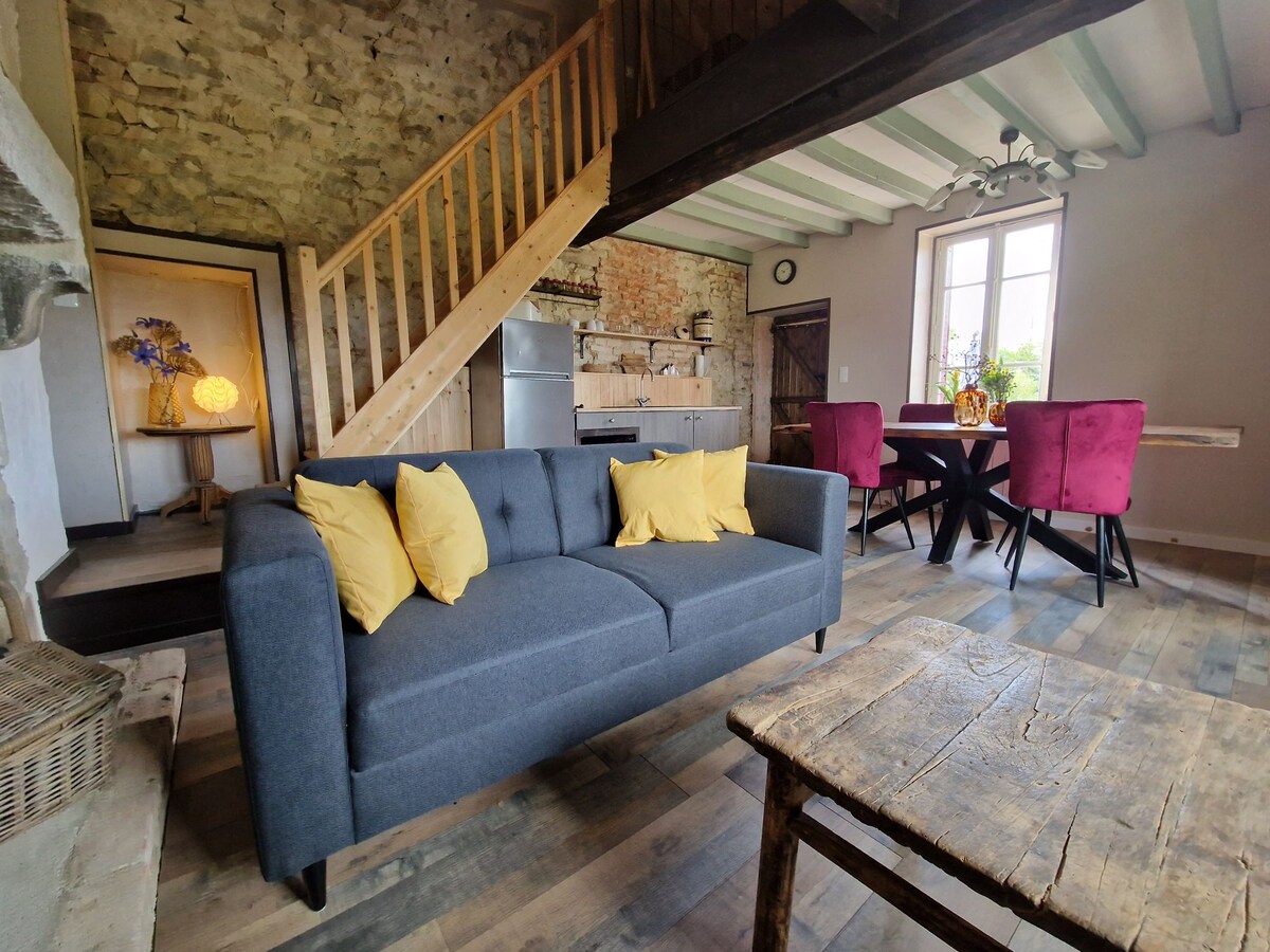 A cozy living space features a gray sofa adorned with yellow cushions, inviting relaxation. A rustic wooden table is positioned nearby, complemented by purple dining chairs. The kitchen area is visible with modern appliances, and natural light filters in through a window framed by wooden beams.