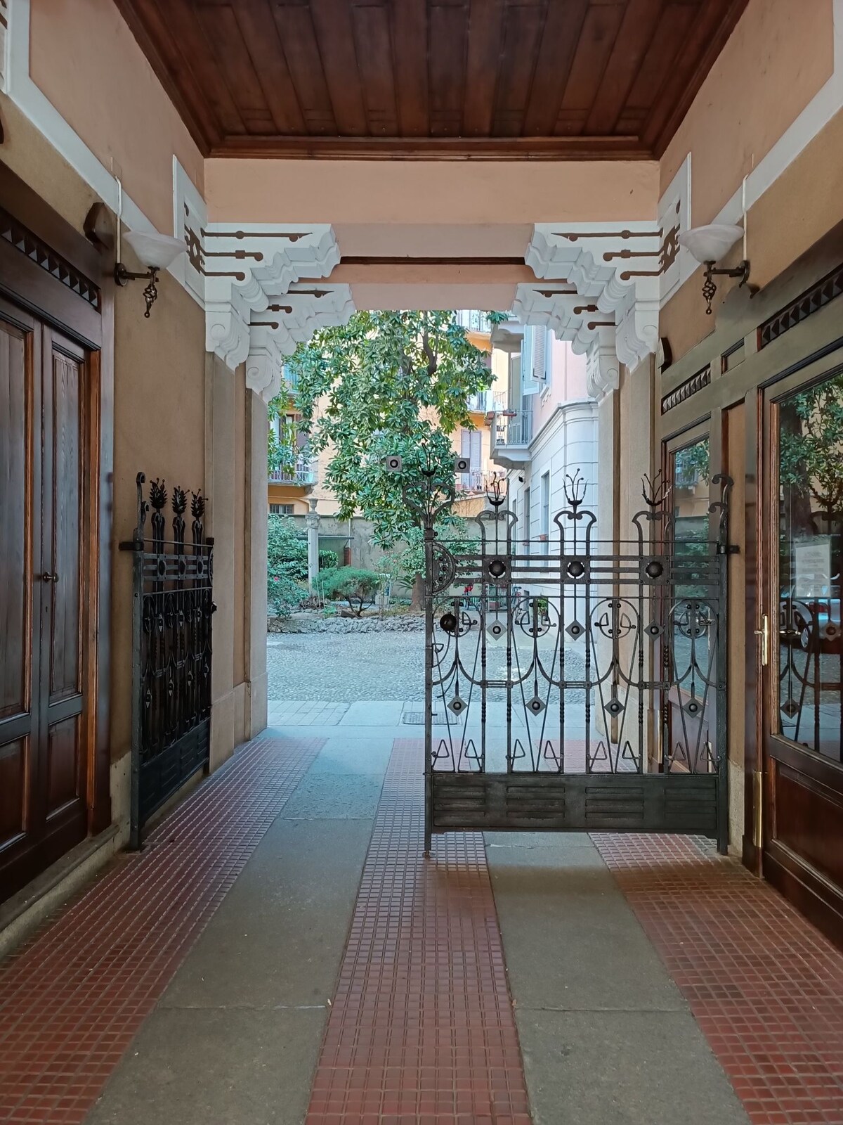 An elegantly designed entrance features intricate wrought iron gates and wooden doors. Soft light filters through the arched passageway, revealing a glimpse of a tranquil courtyard adorned with greenery. The combination of decorative elements creates a welcoming yet refined atmosphere.