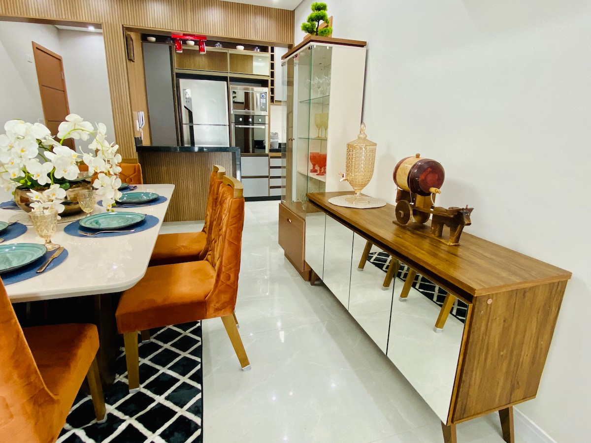 A dining area features a rectangular table set for six, complemented by orange upholstered chairs. A sideboard displays decorative items, including a glass vase and a small sculpture. The space is illuminated by natural light, with a modern kitchen visible in the background.