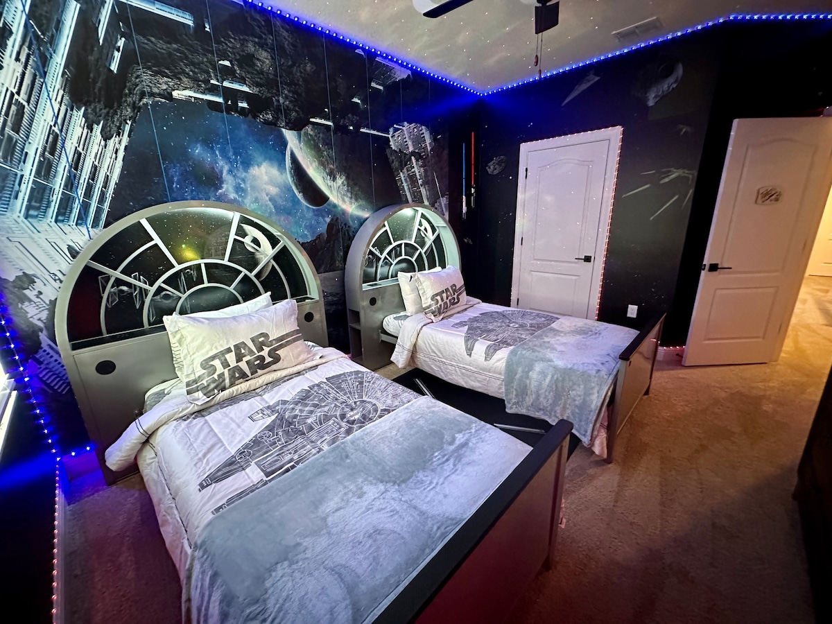 The Star Wars-themed room features two twin beds with sleek headboards and bedding adorned with Star Wars graphics. Neon LED lights run along the ceiling, enhancing the space's celestial ambiance. Dark, themed wallpaper depicting a galactic scene adds to the immersive atmosphere.