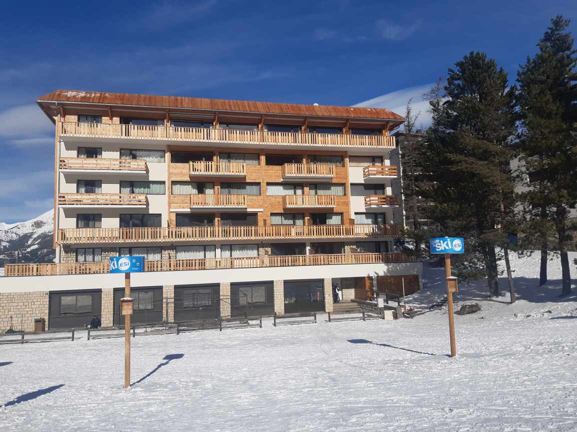 A multi-story building is set amidst snow-covered terrain, featuring balconies on the upper floors. The ground level includes a covered area, while ski signage and a pristine winter landscape add to the ambiance. Pine trees are visible in the background.