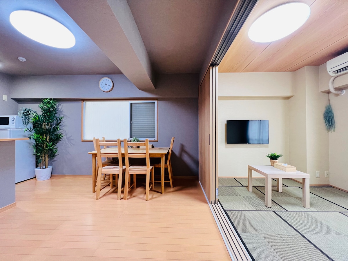 A spacious dining area features a wooden table with six chairs, complemented by a window allowing natural light. A cozy living space is visible with a flat-screen TV mounted on a neutral wall, while traditional tatami mats cover the floor.