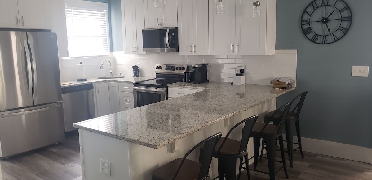 A modern kitchen is presented, featuring stainless steel appliances, including a refrigerator and microwave. A large island with seating for four is highlighted by a sparkling granite countertop. Natural light enters through a window, while a wall clock adds a touch of decor.