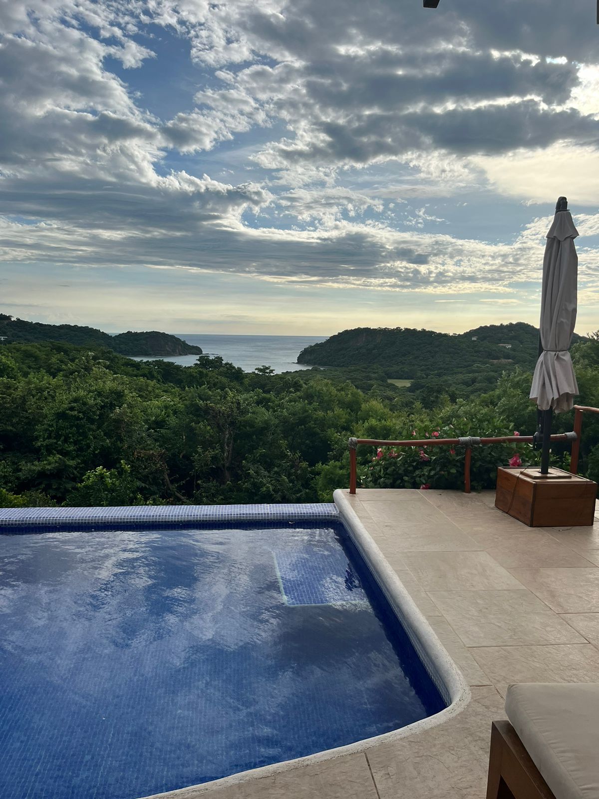 A tranquil pool is situated next to an expansive outdoor area, offering views of lush greenery and the distant Pacific Ocean. A large umbrella provides shade near the water's edge, while dramatic clouds fill the sky above, creating a serene backdrop.