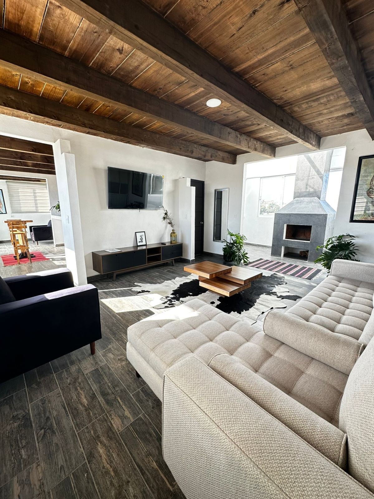 A bright living area is characterized by exposed wooden beams and large windows that allow natural light to fill the space. A light-colored sectional sofa faces a modern fireplace, while a coffee table sits on a soft area rug. Green plants add a touch of nature.