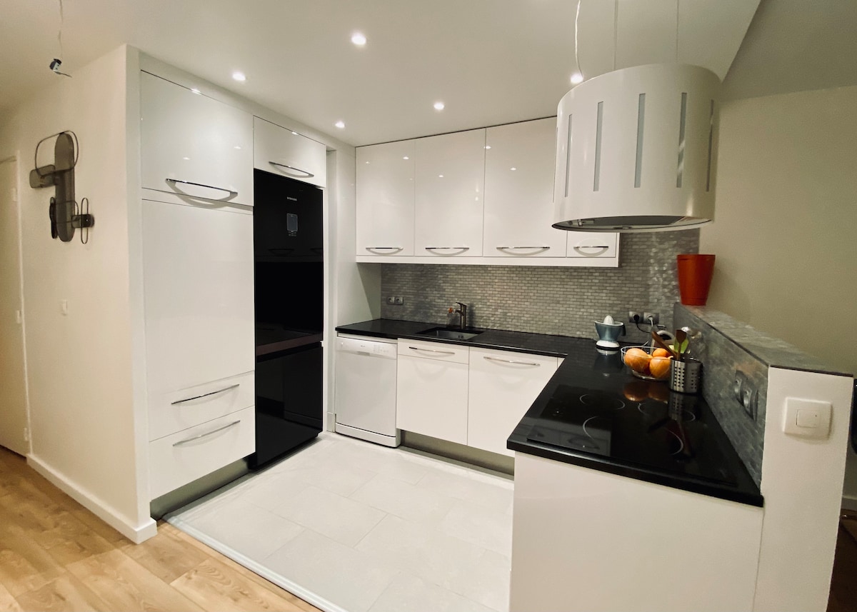 The kitchen features sleek white cabinetry paired with a black countertop. Modern appliances are integrated seamlessly, including a refrigerator and dishwasher. A stylish pendant light hangs above the island. Decorative bowls of fruit add a pop of color to the countertop, enhancing the space's functionality.
