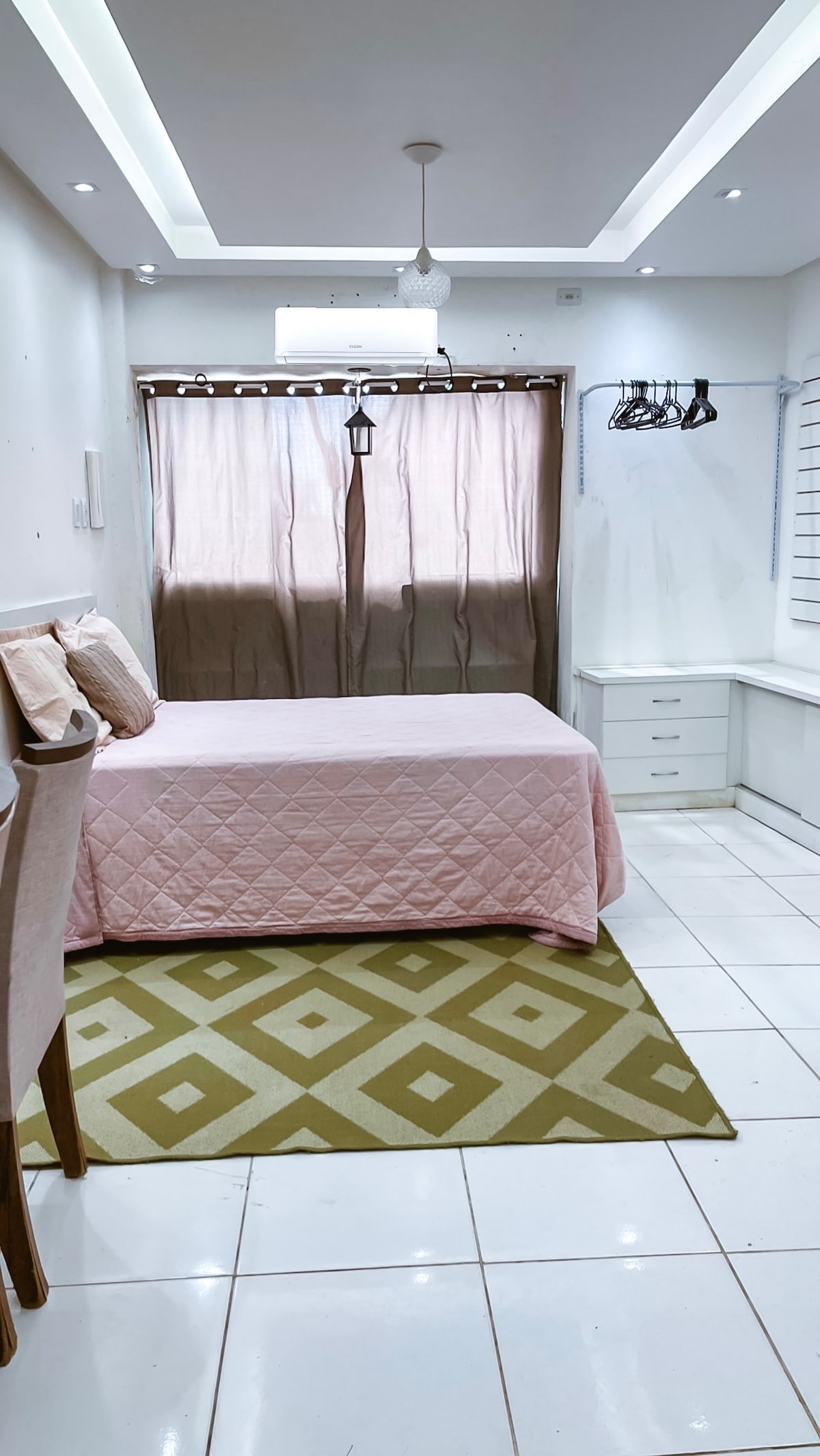 The room features a spacious bed covered with a light pink quilt. A patterned rug lies on the tiled floor, complementing the neutral decor. Natural light enters through a large window covered by brown curtains, while a clothes rack and dresser provide storage options.