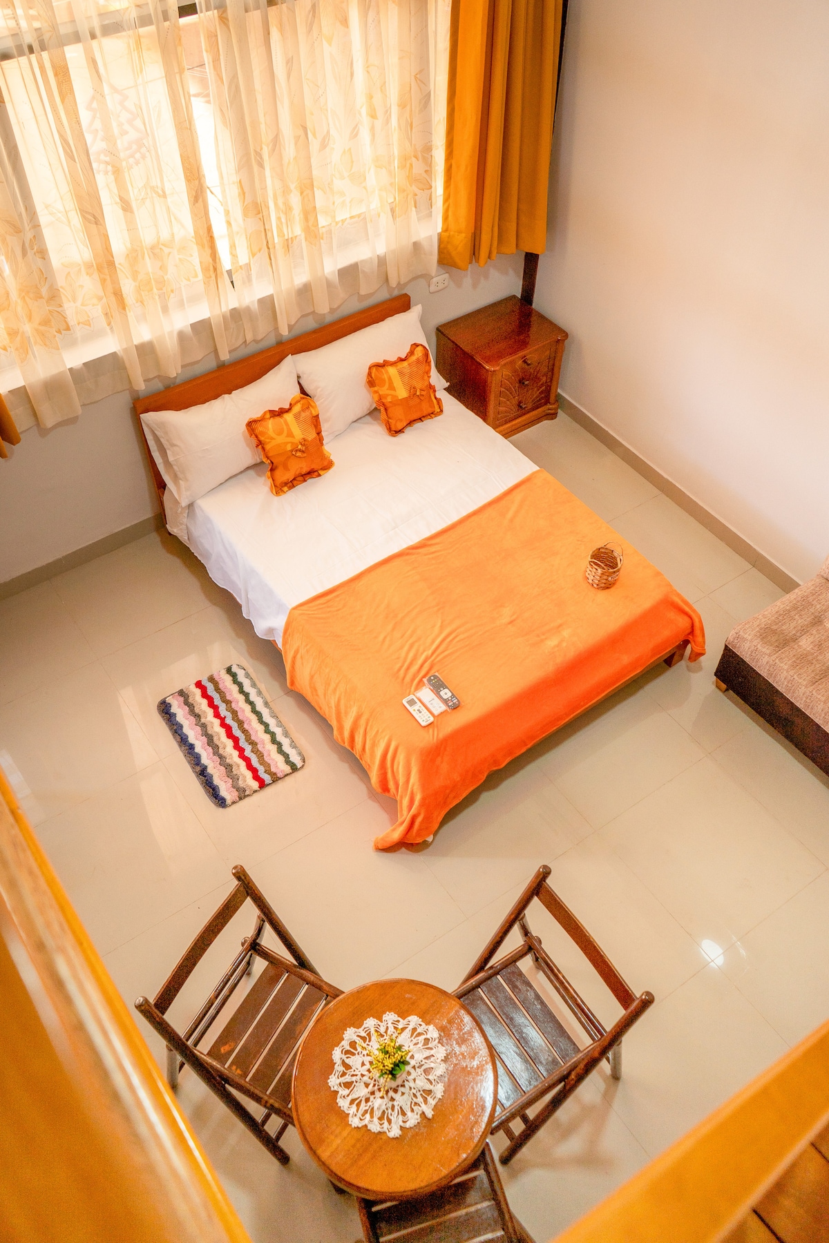 A well-lit sleeping area features a spacious bed with an orange blanket and decorative pillows. Nearby, a small wooden table and two chairs provide a cozy dining space. A colorful rug adds texture to the floor, complemented by large windows with light-filtering curtains.