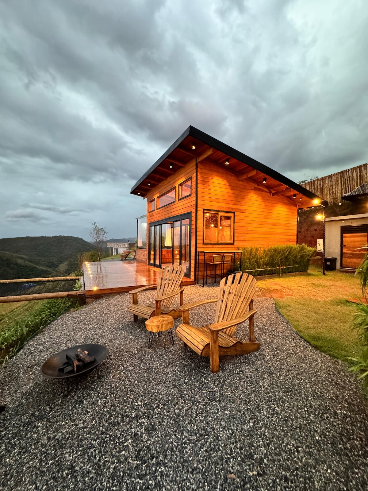 The exterior of a wooden cabin is shown, featuring large glass windows framed by warm wood tones. Two rustic chairs are positioned in front of a fire pit, with a gravel pathway leading to the expansive view of rolling hills and a cloudy sky.