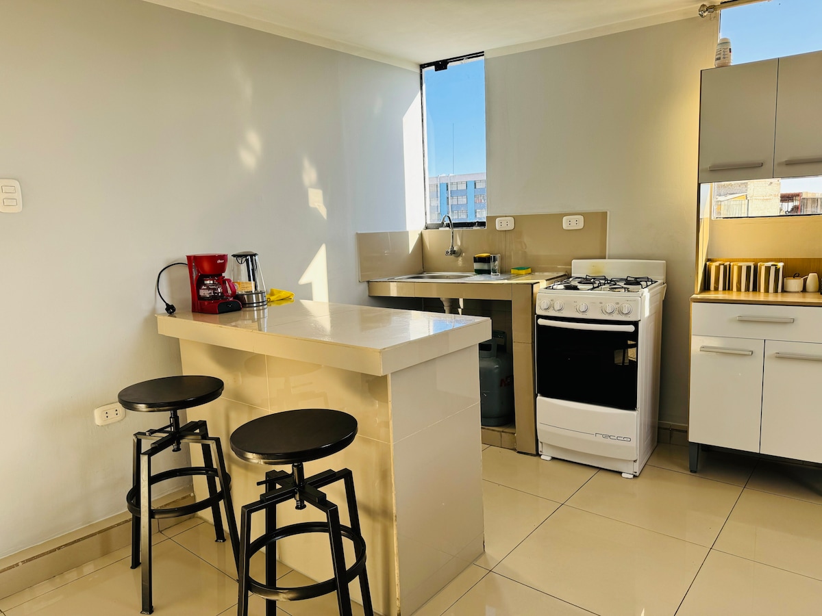 A modern kitchen features a light-colored countertop with two black stools positioned beside it. A red coffee maker and a gas stove are visible, complemented by cabinets and a window providing natural light.