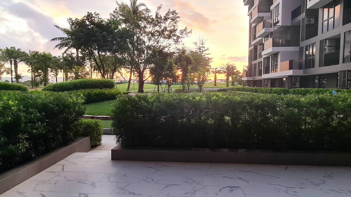 A view from a private terrace showcases manicured greenery framed by lush bushes. The background features a vibrant sunset over the horizon, complementing the serene landscape of the surrounding grounds and palm trees.
