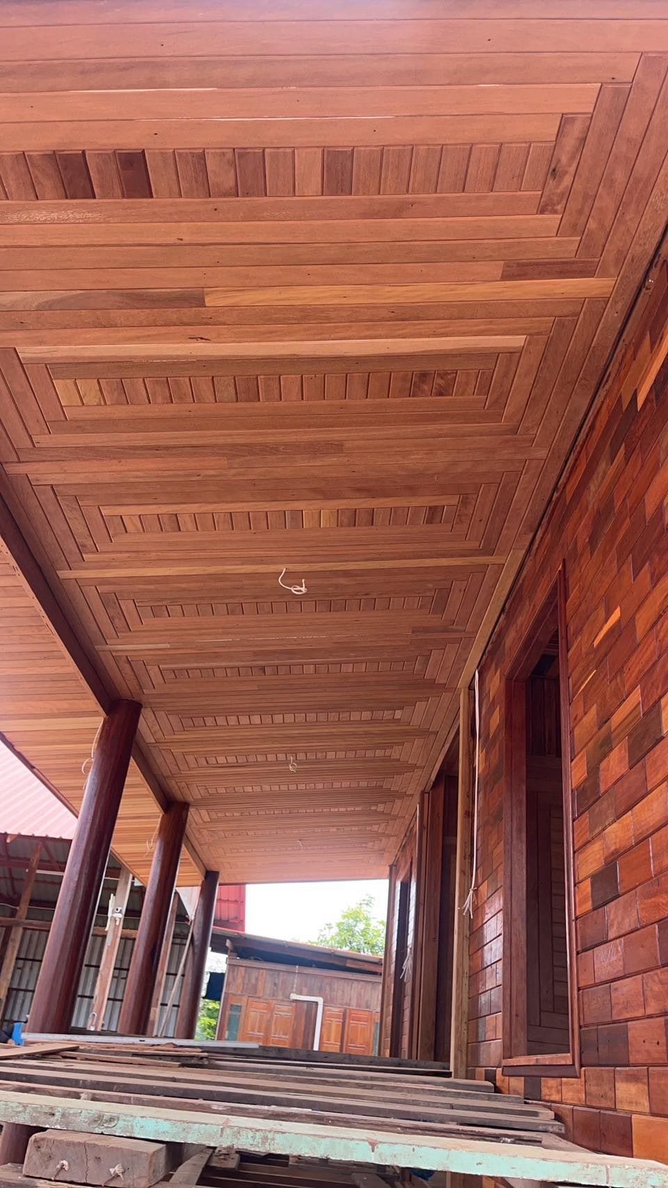 The image captures the underside of a wooden porch, showcasing intricately patterned wooden panels and sturdy columns. The warm tones of the timber contrast with the red brick wall in the background, creating a harmonious blend of natural materials.