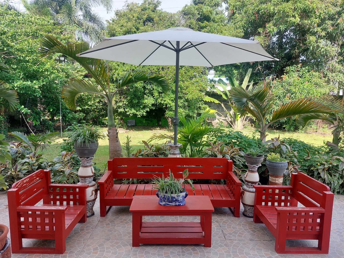 A cozy outdoor seating area is furnished with red wooden chairs and a table, shaded by a large umbrella. The surrounding garden features lush greenery and potted plants, creating a peaceful environment for relaxation.
