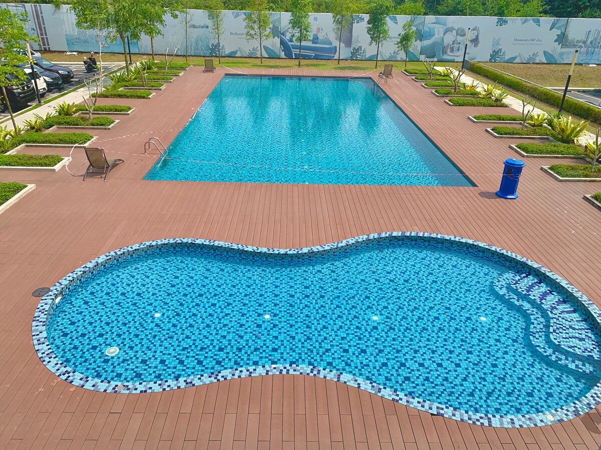 A large swimming pool is situated within a landscaped area, featuring a distinct free-form shape. A secondary rectangular pool is positioned alongside, surrounded by smooth tiles and neatly arranged greenery. Several lounge chairs are placed comfortably around the pool for relaxation.