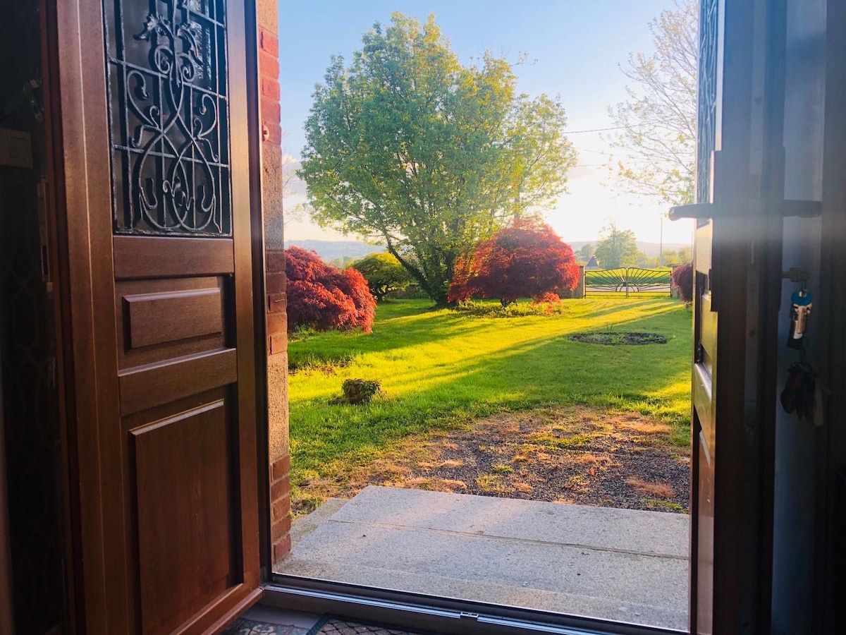 The entrance door is open, revealing a lush garden with vibrant green grass and a variety of trees, including a red maple. Soft sunlight filters through, highlighting the serene outdoor space that invites moments of relaxation.