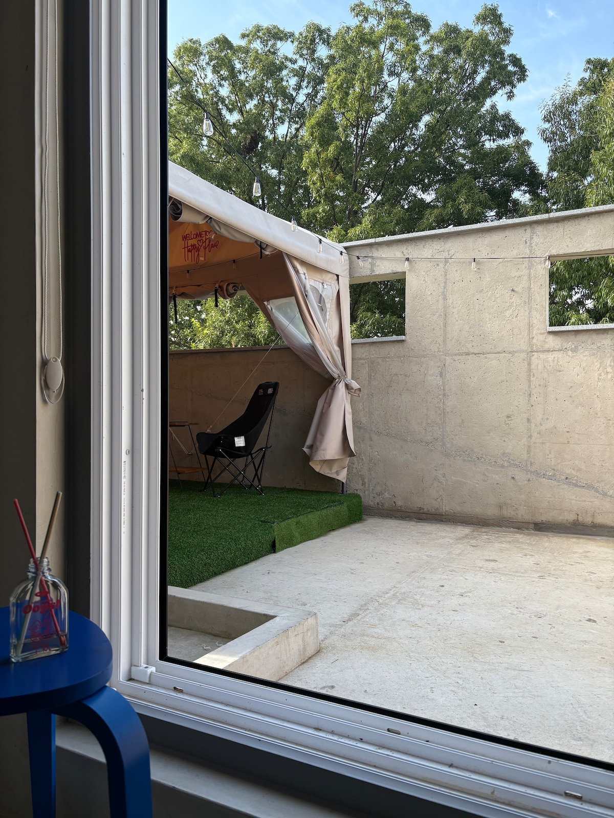 A view from a window showcases a spacious outdoor area featuring a tent and a black folding chair. The tent is adorned with soft draping, and green artificial grass is positioned beneath, creating a relaxed atmosphere in the garden setting.