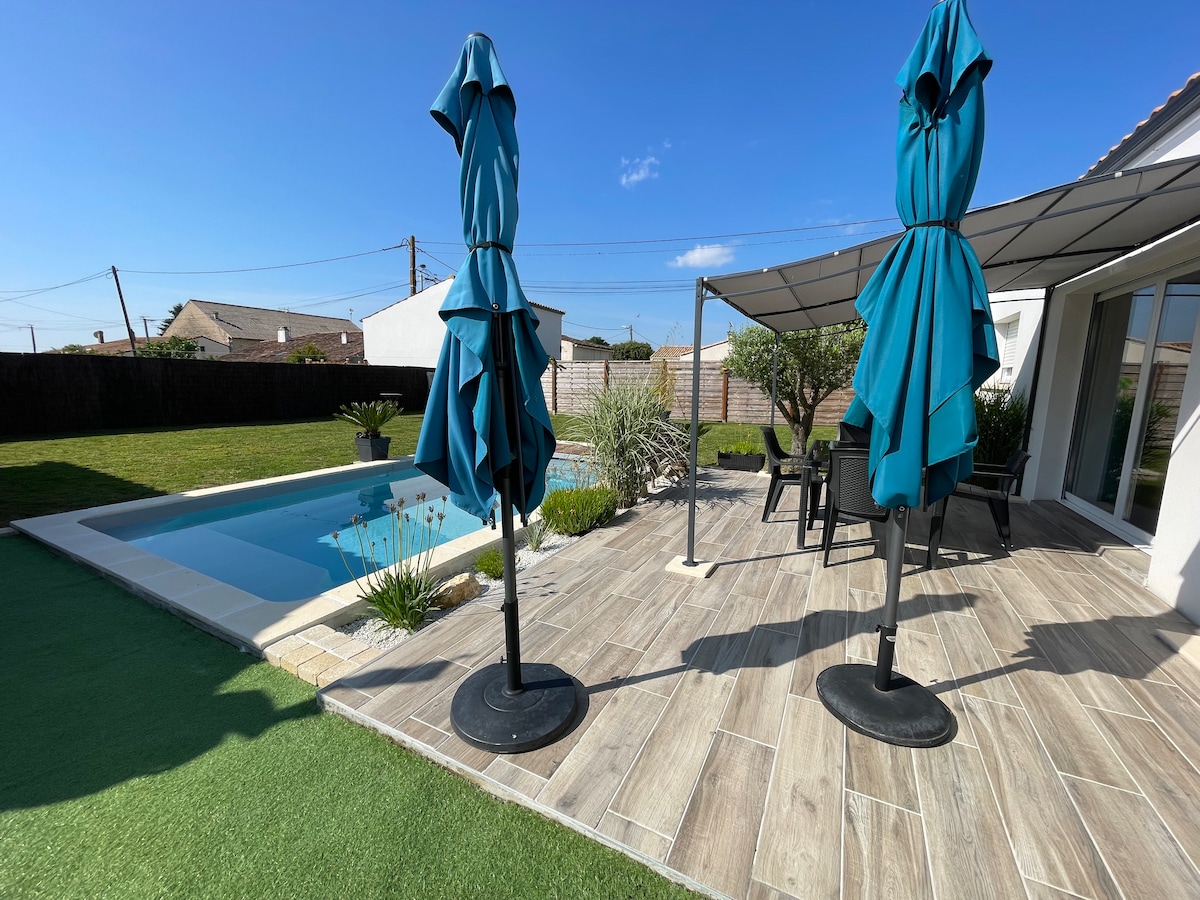 A serene outdoor area is showcased, featuring a pool surrounded by stone flooring and greenery. Two blue umbrellas stand on either side, providing shade over a seating arrangement. The sunlit area indicates a relaxing space perfect for enjoying the garden view.