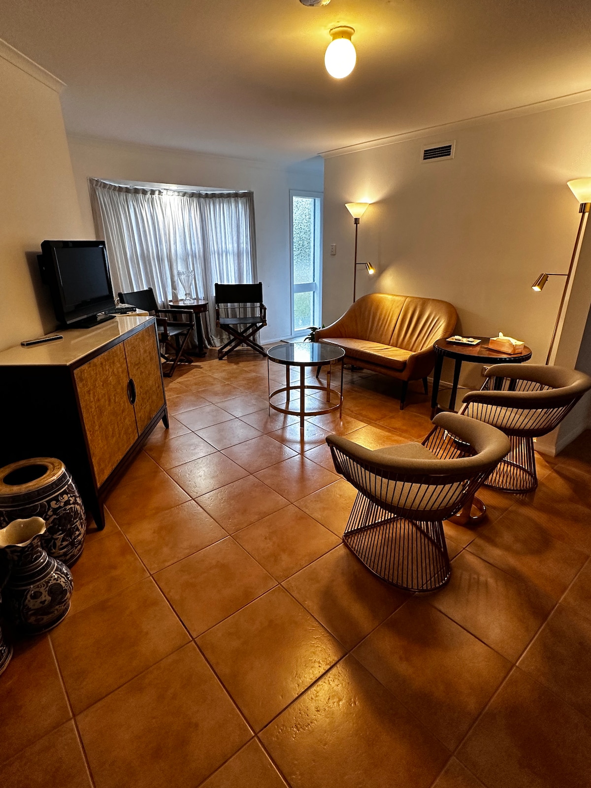 A welcoming living area features a comfortable brown upholstered sofa paired with two modern wire chairs. A glass coffee table sits in the center, and a television is positioned atop a wooden cabinet. Natural light filters through sheer curtains, creating a relaxed ambiance.