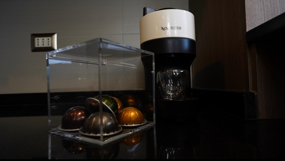 A modern coffee machine is positioned on a dark countertop next to a transparent container holding coffee capsules in various colors. Behind it, a wall outlet is visible, providing easy access to power.
