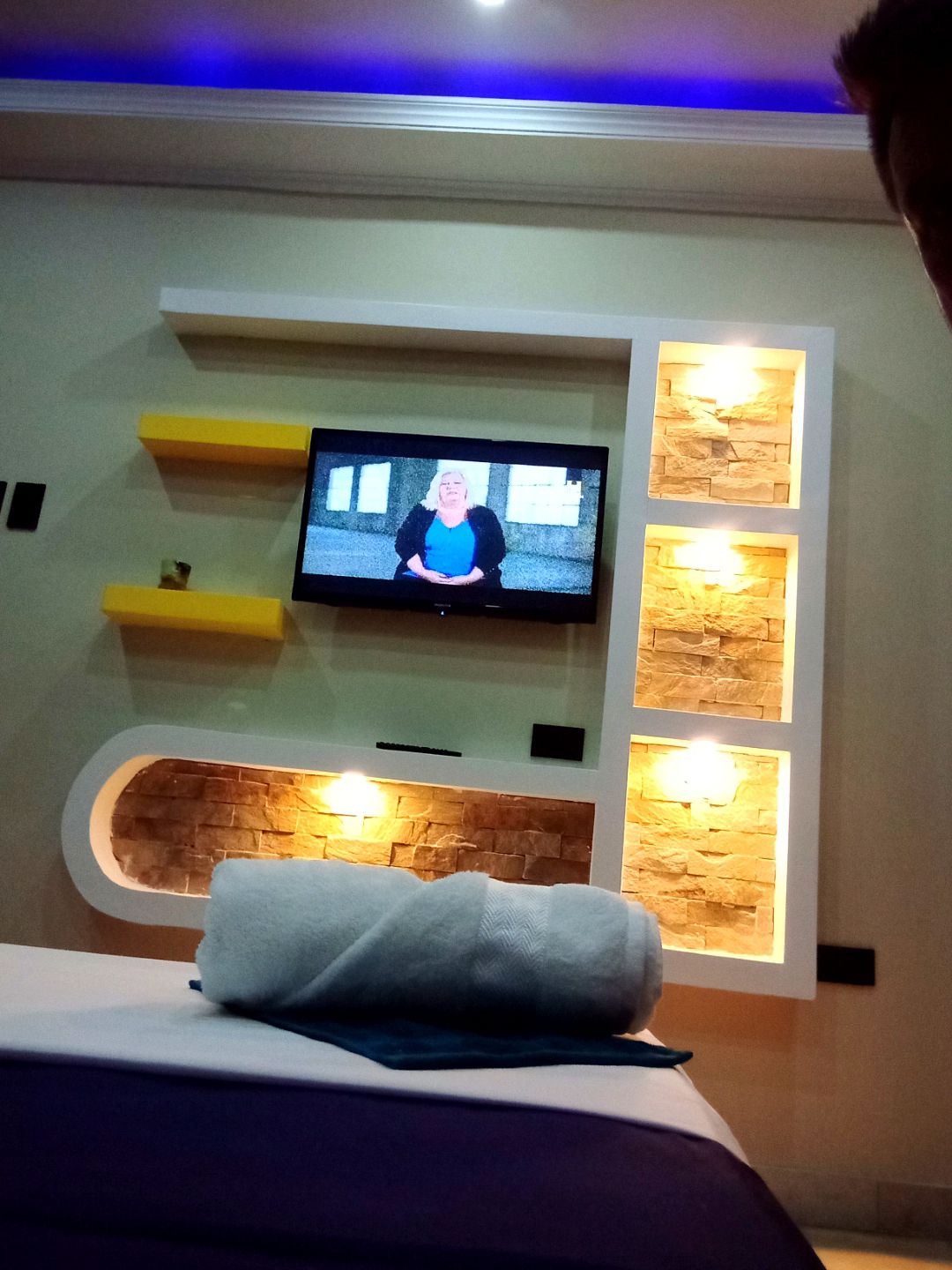 A modern room features a wall-mounted television set against a stylish backdrop of decorative stone and illuminated shelves. A neatly rolled towel is placed on the spacious bed, contributing to a clean and organized atmosphere. Soft lighting enhances the overall ambiance.
