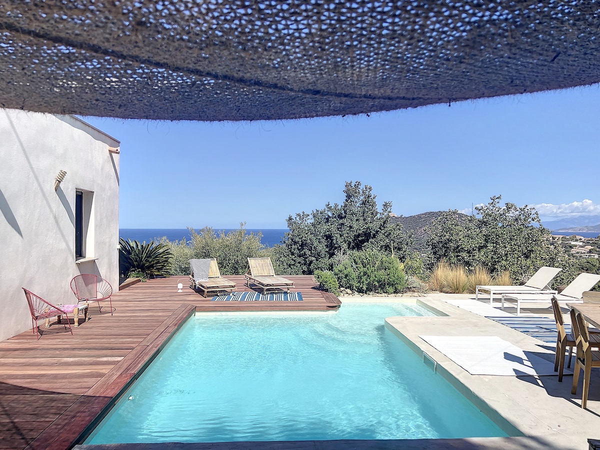 A serene pool area is framed by wooden decking, with sun loungers arranged around the heated pool. Lush greenery and distant hills are visible in the background, while a clear blue sky reflects above the water. A shaded area provides relief from the sun.