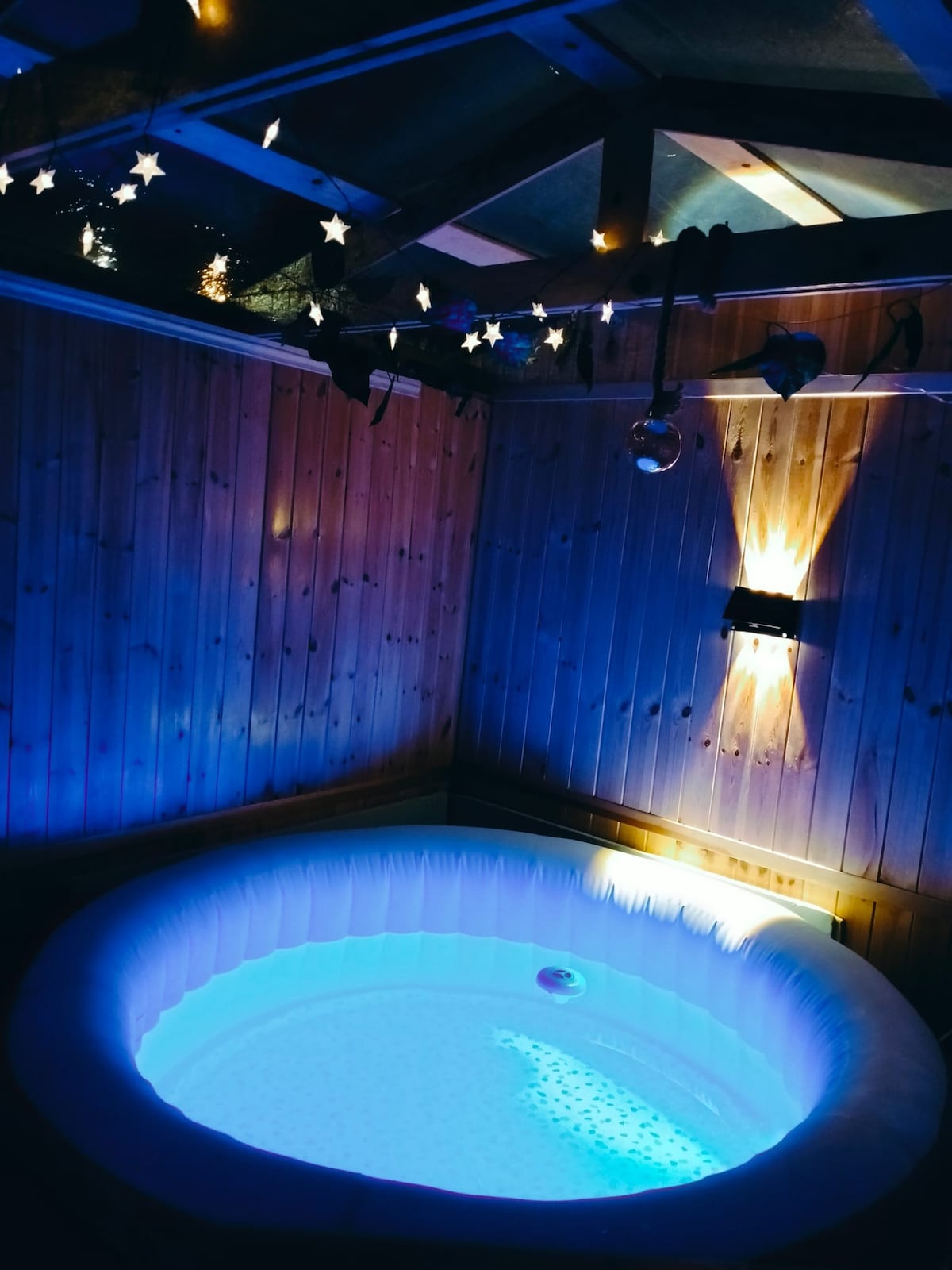 A heated jacuzzi is located in a cozy wooden enclosure, illuminated by ambient lighting and decorative stars hanging from the ceiling. Soft blue light reflects off the water, creating a relaxing atmosphere in the space.