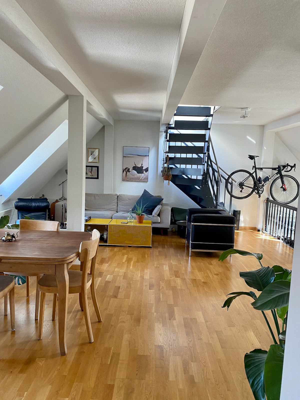 A bright and airy living space is shown, featuring hardwood floors and large windows. A dining table is positioned in the foreground, while a cozy seating area includes various seating options. A set of stairs leads to an upper level, and a bicycle is mounted on the wall.