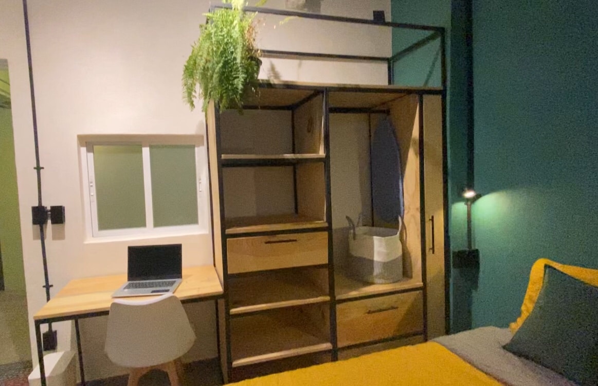 A compact living area features a spacious bed with a yellow throw draped across it. A study desk with a laptop sits beside a modern bookshelf that includes a laundry basket. A small indoor plant adds a touch of greenery above the wardrobe, and a window allows natural light to enter.