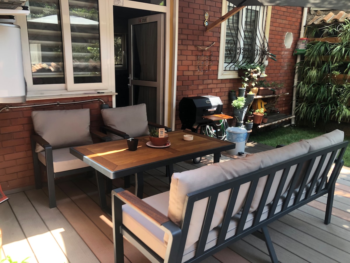 An outdoor seating area features a wooden table surrounded by two cushioned armchairs and a bench. A grill is positioned nearby, while potted plants and greenery enhance the space. Natural light filters through the area, creating a relaxed atmosphere.