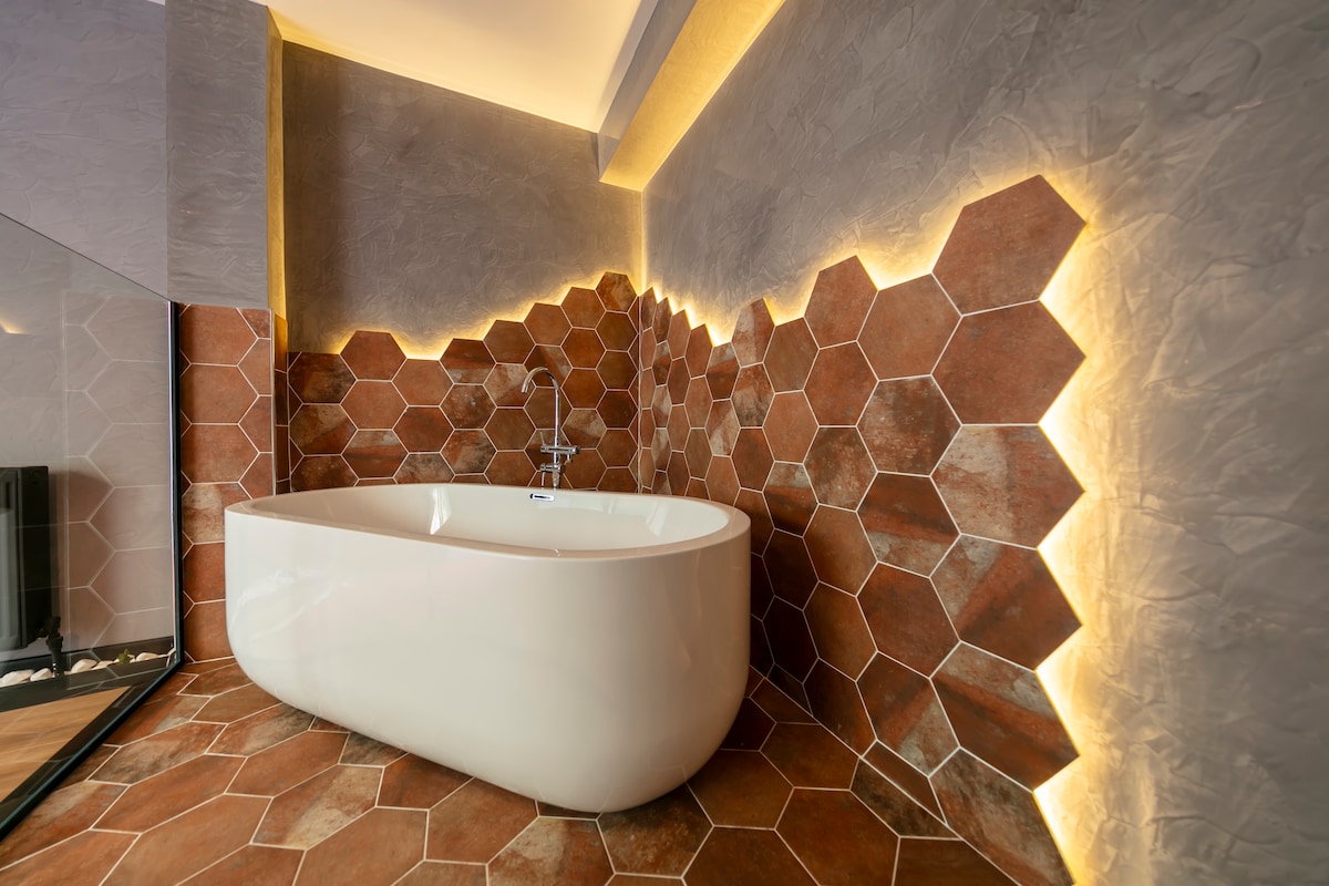 A modern bathroom depicts a freestanding bathtub against a stylish hexagonal tile wall. Soft, warm lighting enhances the unique texture of the tiles, creating a calming and inviting atmosphere. The floor features coordinating tiles that harmonize with the overall design.