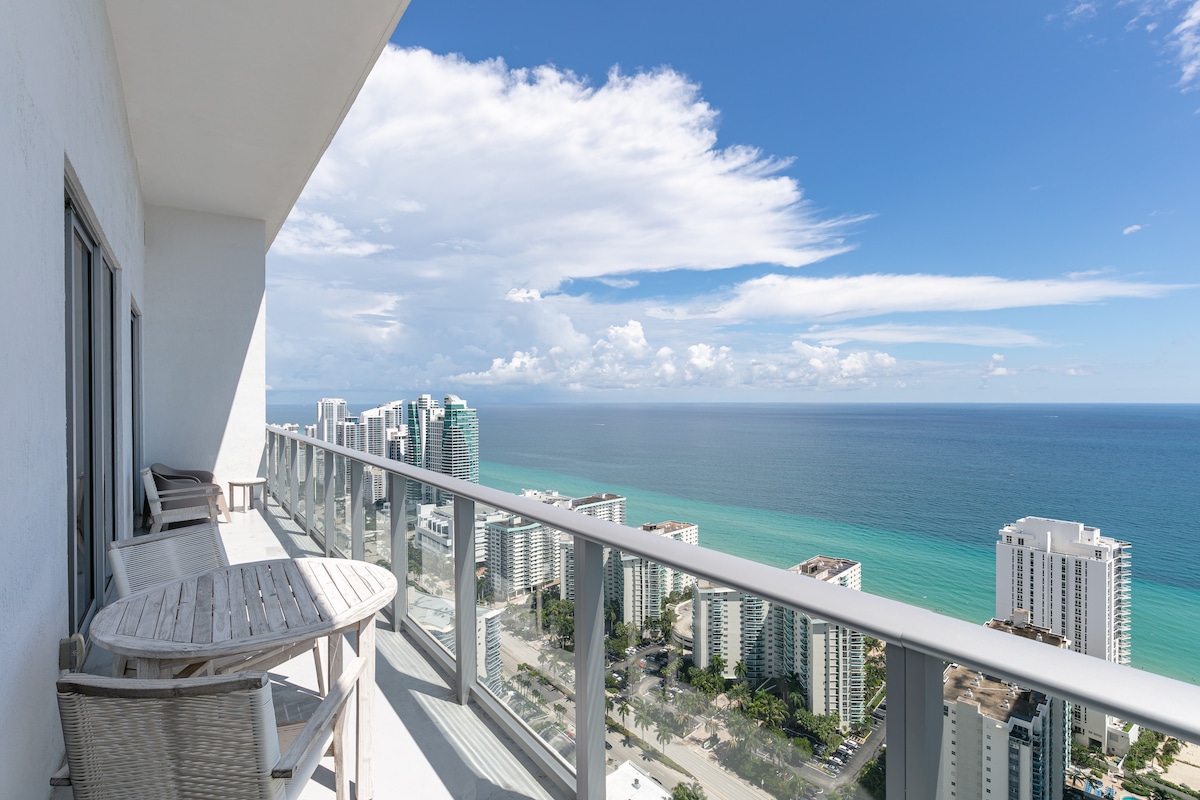 A spacious balcony offers panoramic views of the ocean and cityscape, featuring a wooden table and chairs. The clear blue sky above contrasts with the turquoise water below, creating a serene outdoor space ideal for relaxation.