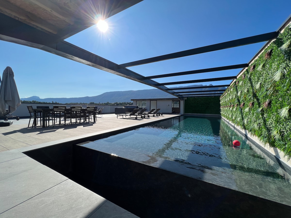 A rooftop pool area is revealed, featuring a modern design with a clear water pool and surrounding sun loungers. A dining table is positioned nearby, with an umbrella and a lush vertical garden providing a vibrant backdrop against the distant mountains under a bright sky.