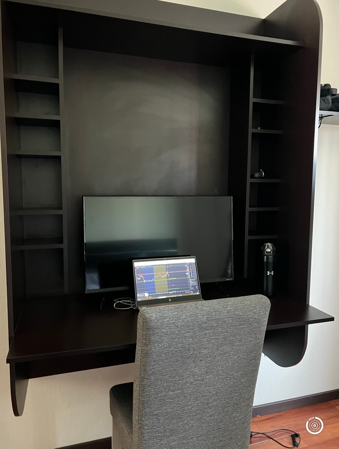 A dedicated workspace is showcased, featuring a large television mounted above a sleek dark desk. A laptop is positioned in front of a comfortable gray chair, with shelves on either side displaying minimal decor. A water bottle rests on the desk, contributing to a functional layout.