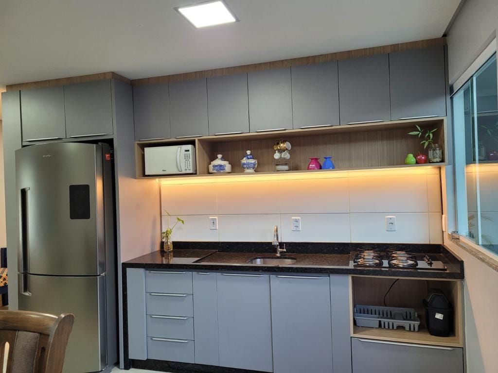 A modern kitchen is equipped with a stainless steel refrigerator, an integrated microwave, and a stovetop with two burners. The countertop is finished in dark granite and features decorative items and plants on display. Under-cabinet lighting illuminates the workspace, enhancing visibility.