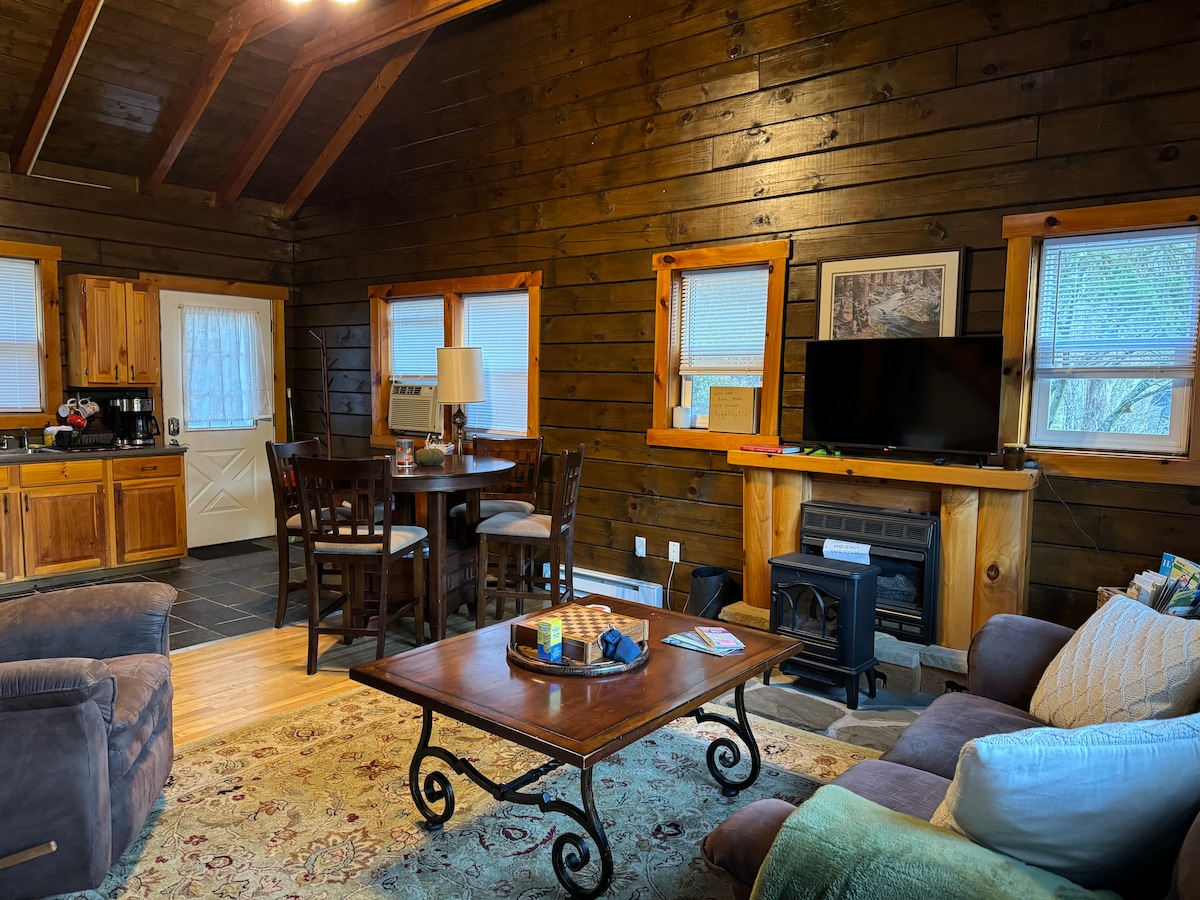 The interior of the cabin features warm wooden walls and a high ceiling, creating a cozy atmosphere. A rustic coffee table is centered in the living area, surrounded by comfortable seating and a dining table. Large windows allow natural light to illuminate the space.