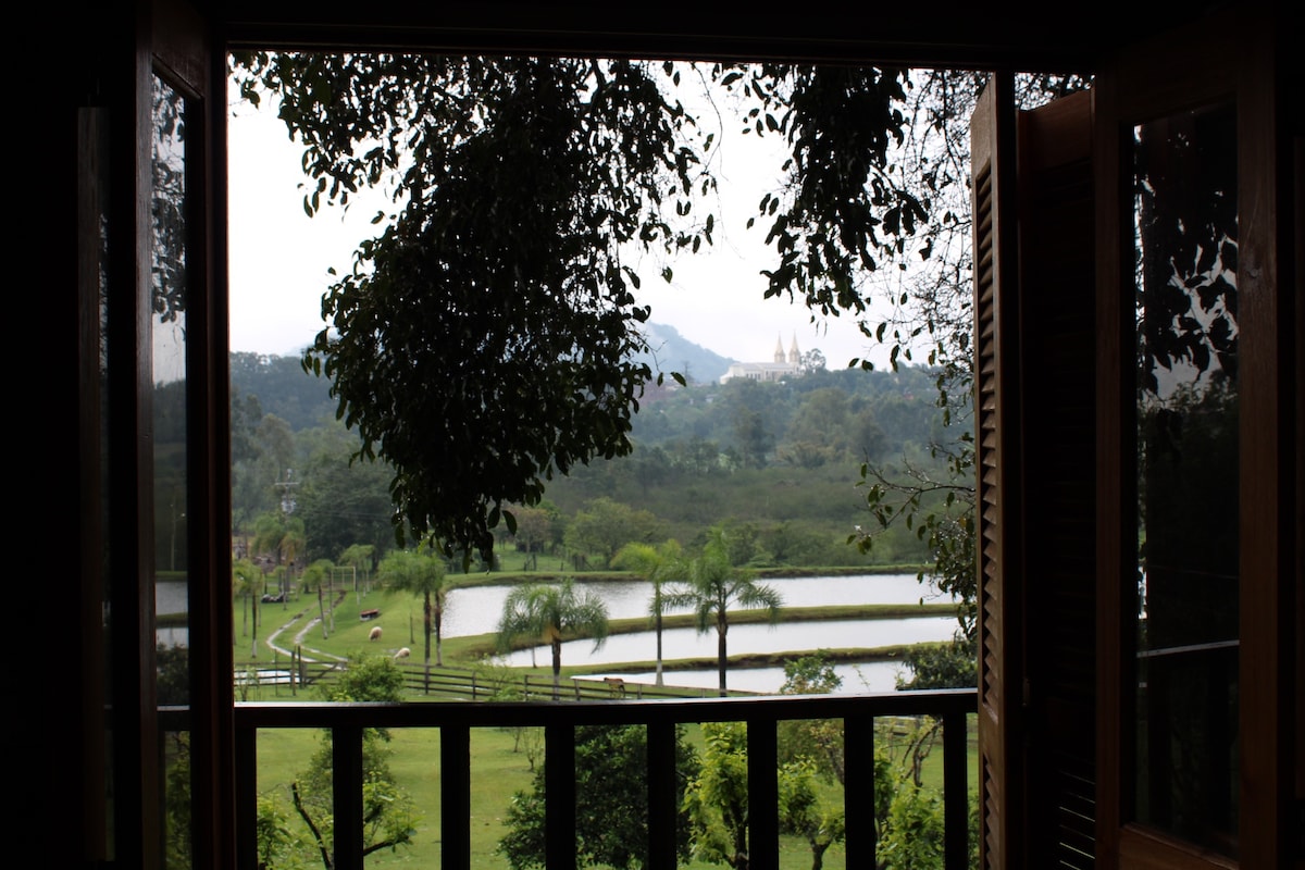 A view is provided from the open window, showcasing a lush landscape with various trees and a serene lake. The distant outline of a church is visible, framed by greenery and hills in the background.