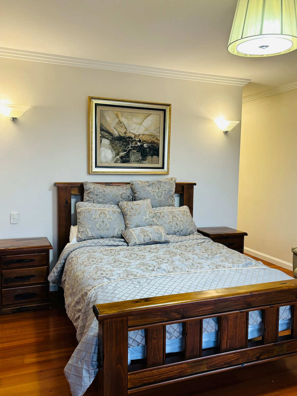 A cozy bedroom features a wooden bed frame with a patterned comforter and several decorative pillows. Soft lighting is provided by wall sconces and a pendant light above. A framed piece of art hangs above the bed, complementing the warm tones of the wooden furniture.