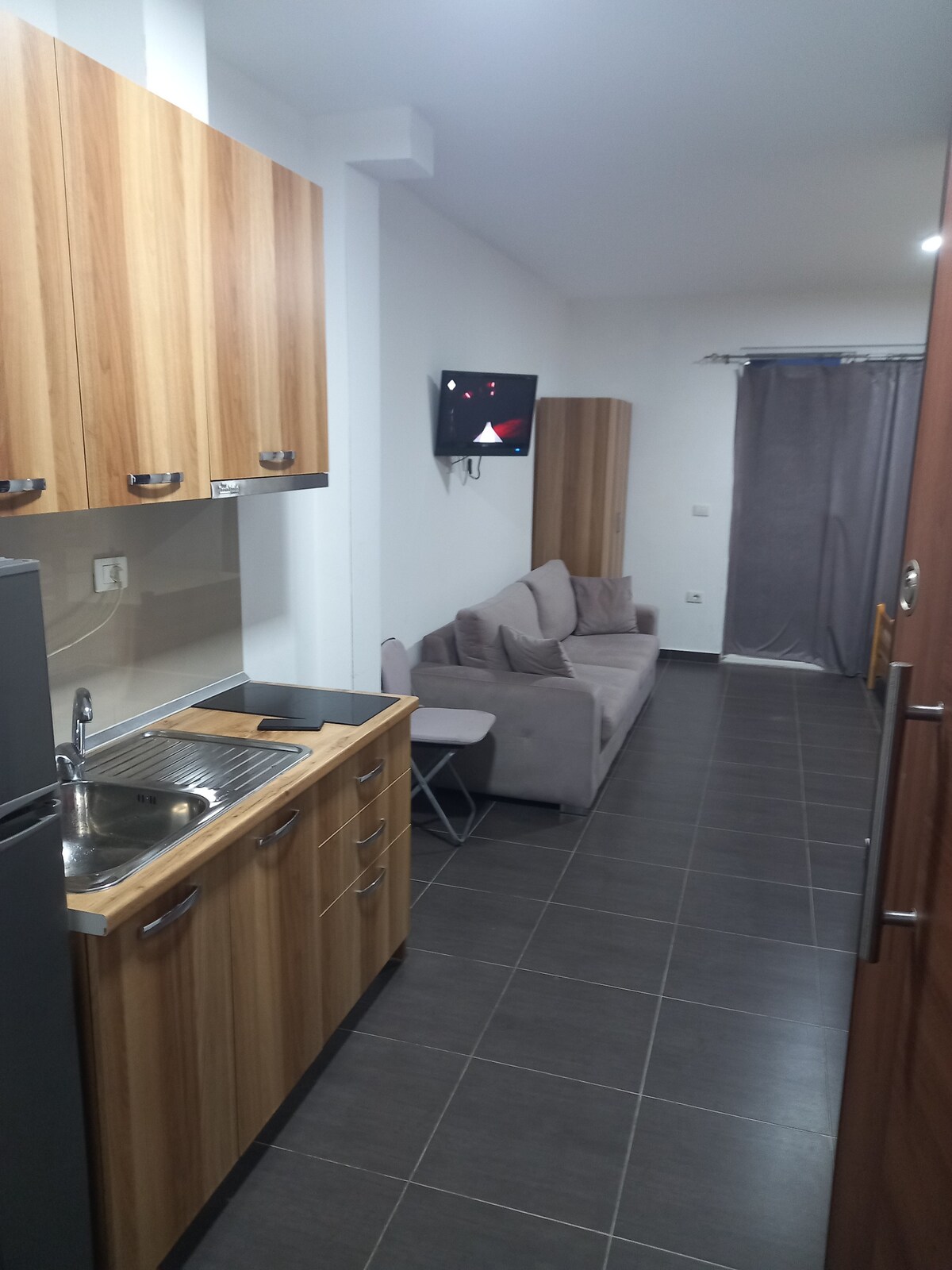 A modern kitchenette is visible, featuring wooden cabinets and a stainless steel sink. A compact dining area includes a small table and chair. The living space is furnished with a gray sofa, and a television is mounted on the wall, illuminated by soft, natural light from nearby windows.